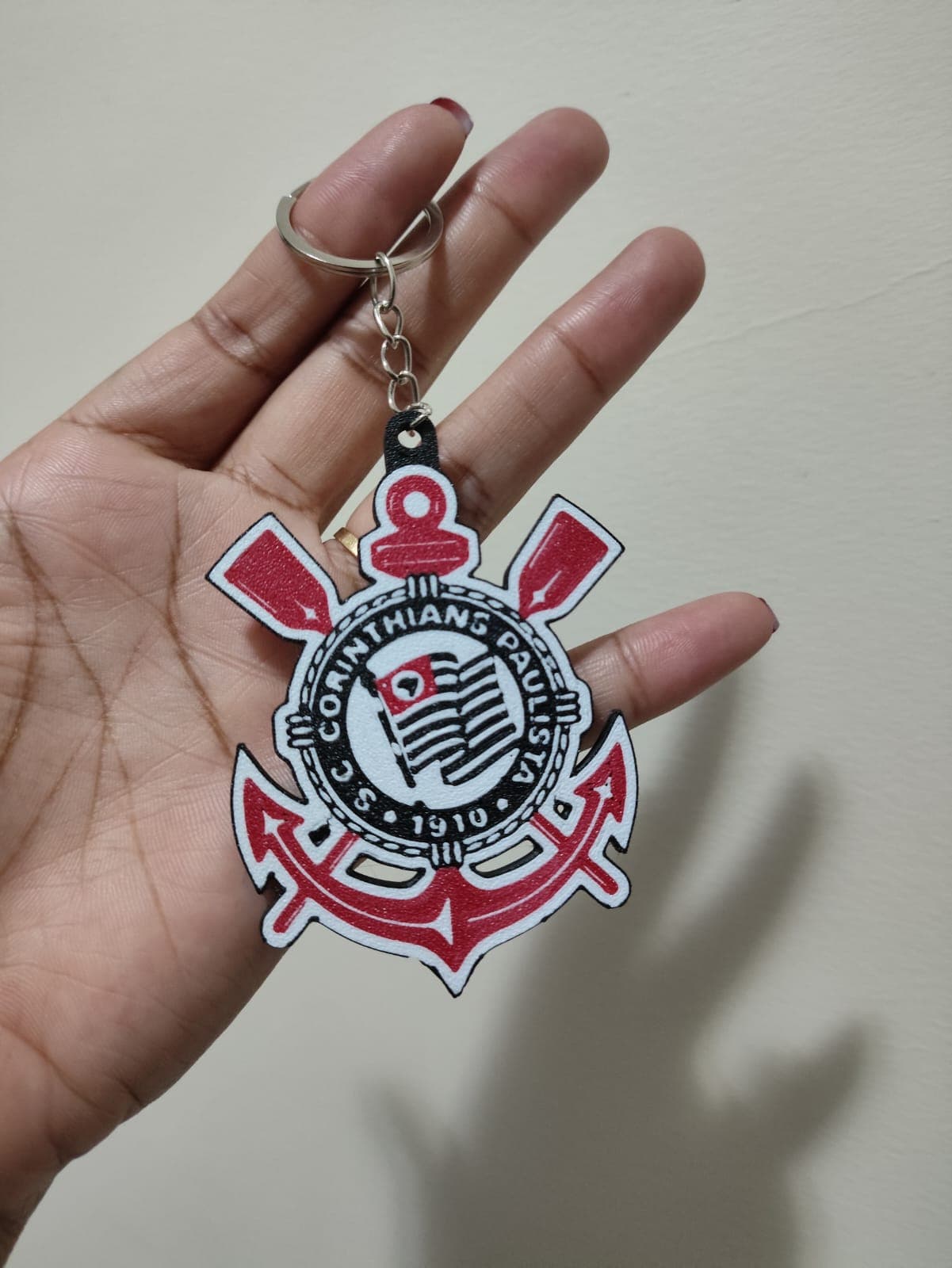 Mão segurando chaveiro com escudo do Corinthians em vermelho, preto e branco.