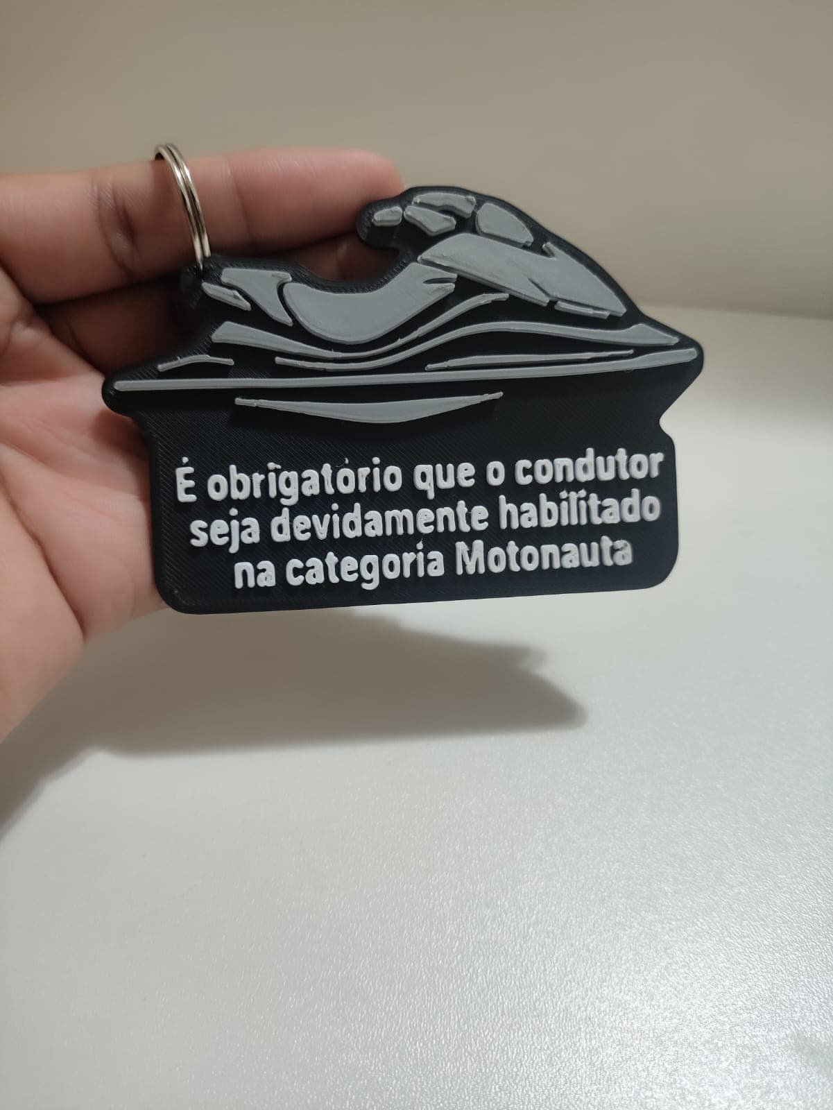 Mão segurando chaveiro preto com jet ski em relevo e texto branco sobre motonauta.