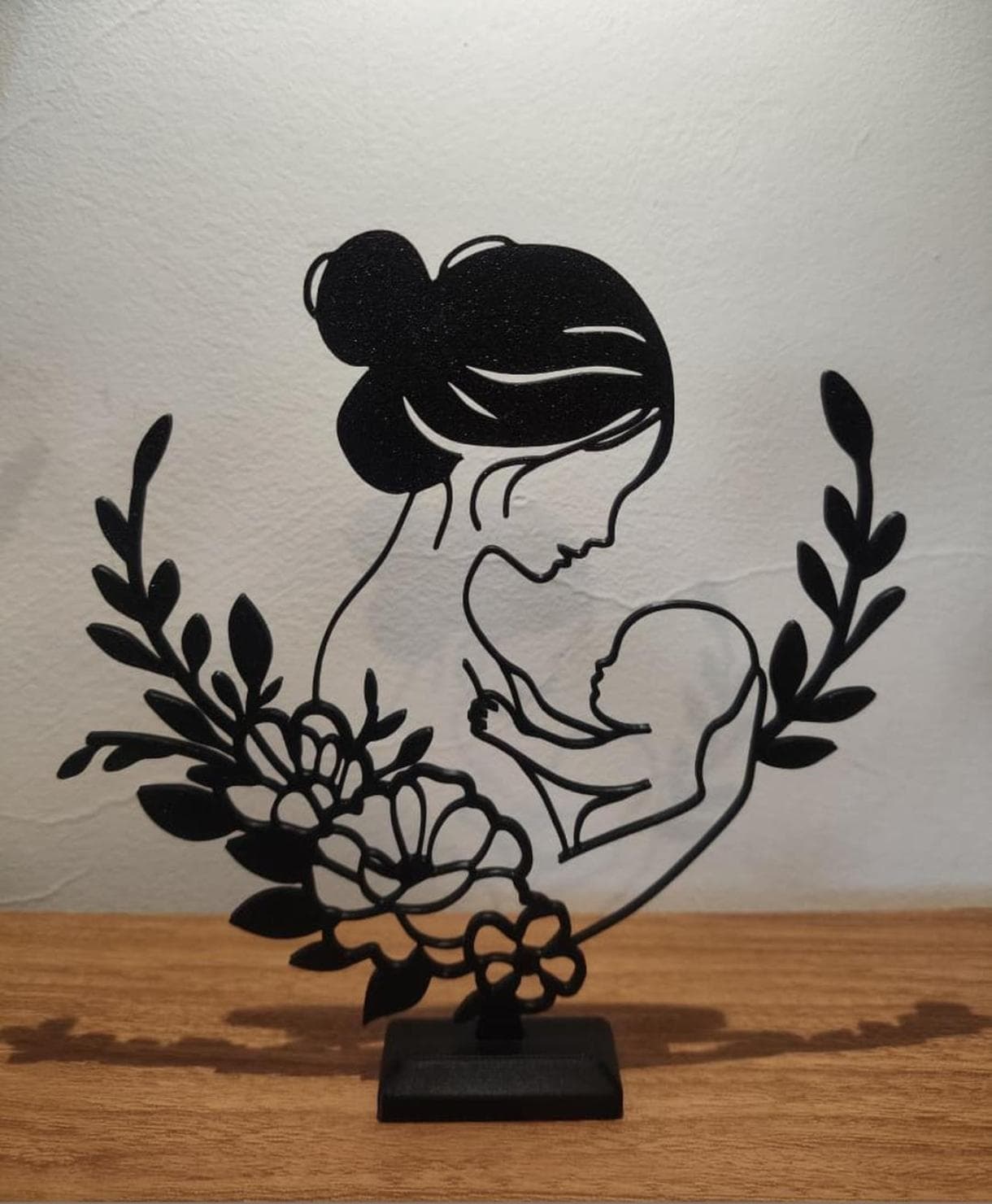 Escultura em silhueta preta com mãe, bebê e flores, sobre madeira clara.
