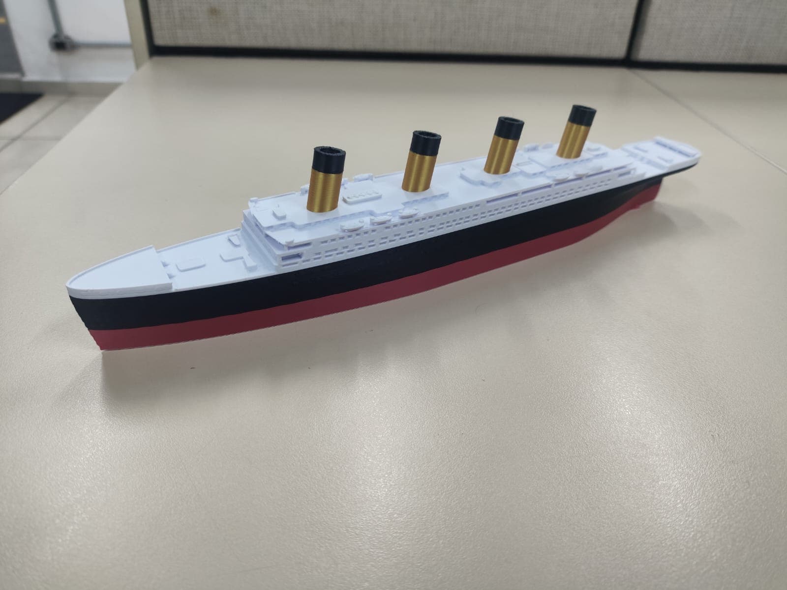 Miniatura impressa em 3D do navio Titanic com casco vermelho, preto e branco e chaminés douradas, sobre superfície clara.