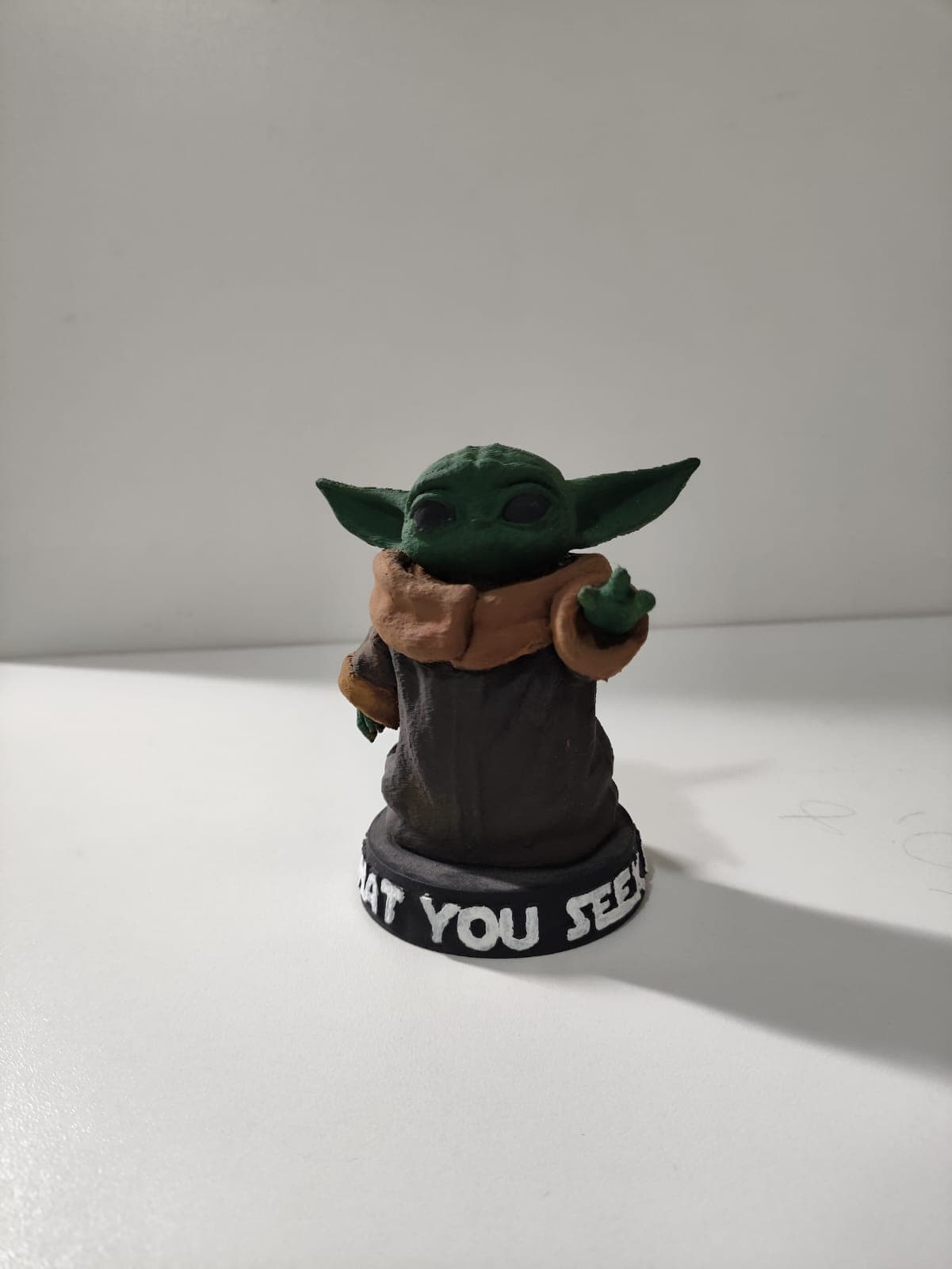 Miniatura de Grogu com mão levantada sobre base preta com texto branco, fundo claro.